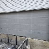 A gray multi-panel garage door installed under white horizontal siding, with a partial metal railing in the foreground.