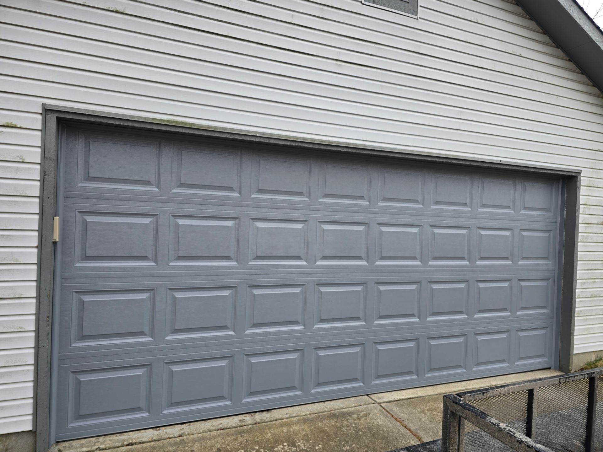 A gray, multi-panel garage door installed on the exterior of a building with white horizontal siding.