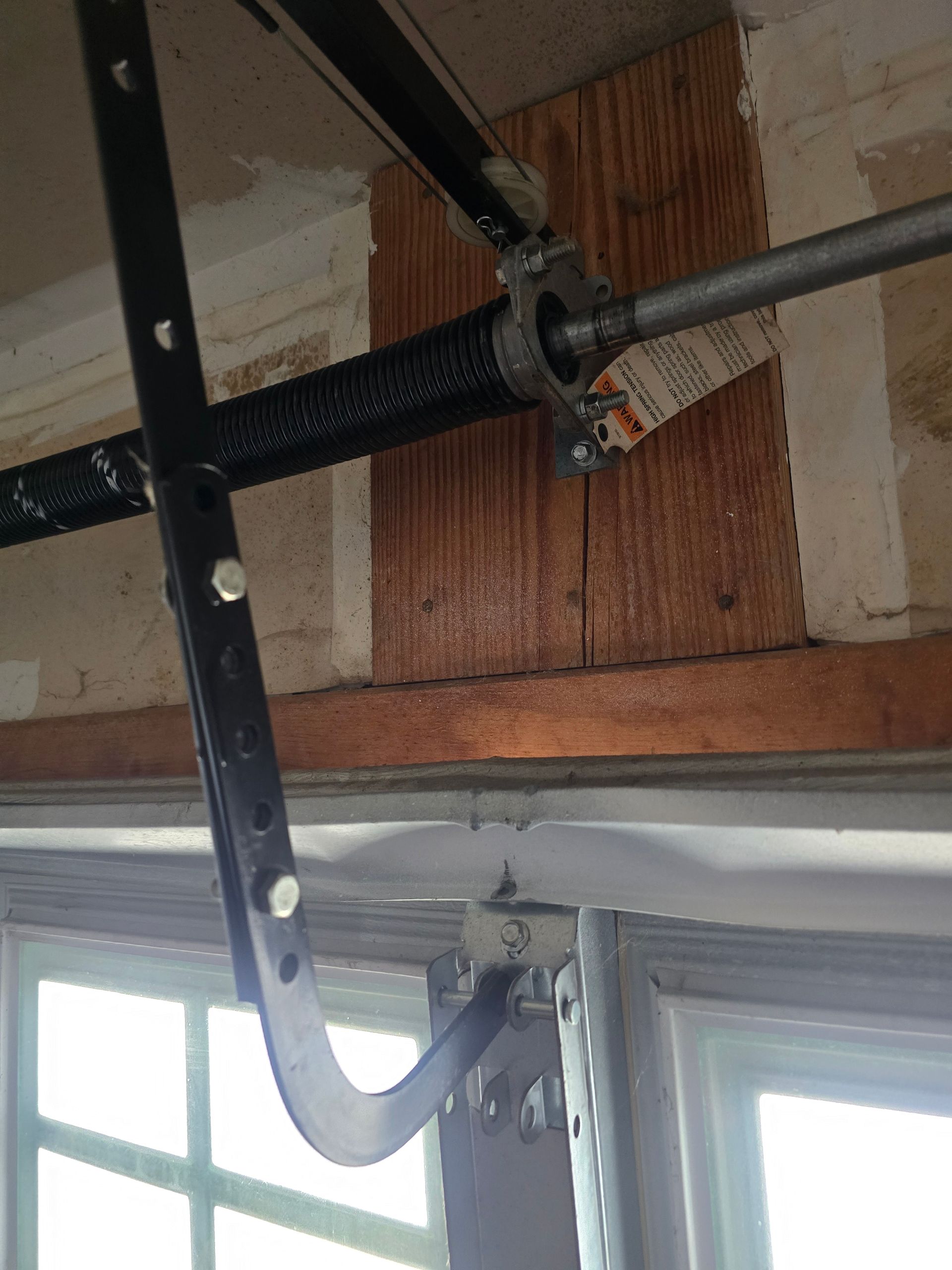 A black metal arm of a garage door opener is attached to the hardware and a torsion spring mounted on a wooden header.