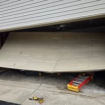 A beige garage door is partially collapsed and sagging in the middle, with a red ladder lying on the concrete floor.