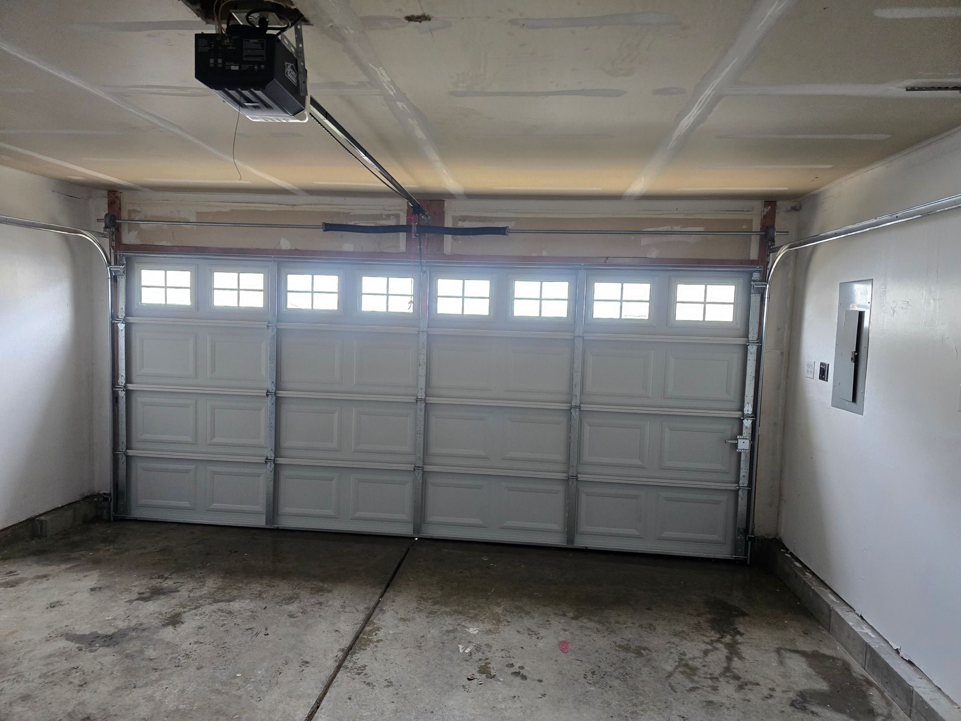 Interior view of a garage featuring a closed white four-panel garage door with windows and a ceiling-mounted opener.
