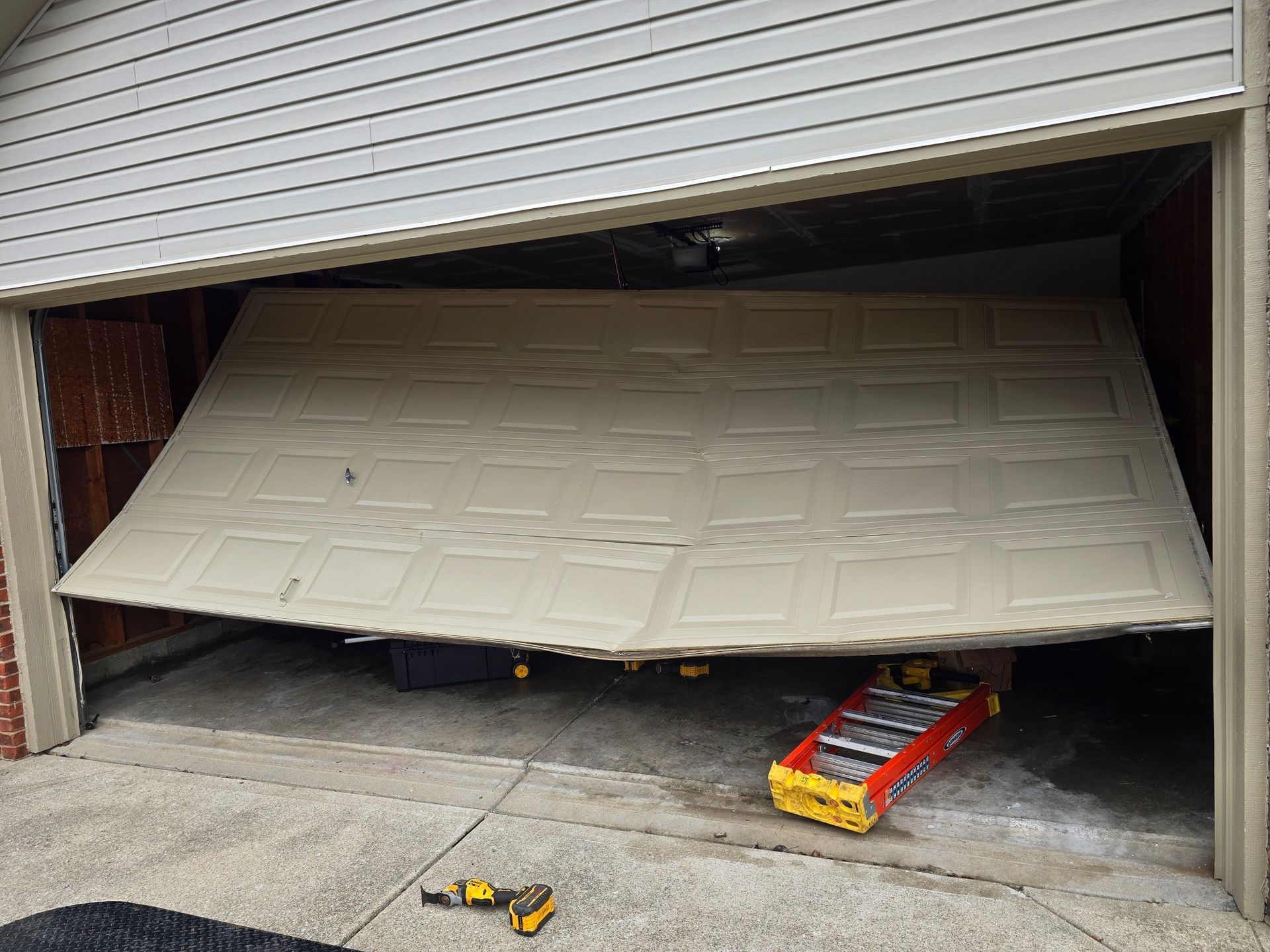 A beige residential garage door hangs crooked and off its track, with a ladder and tools on the concrete floor below.