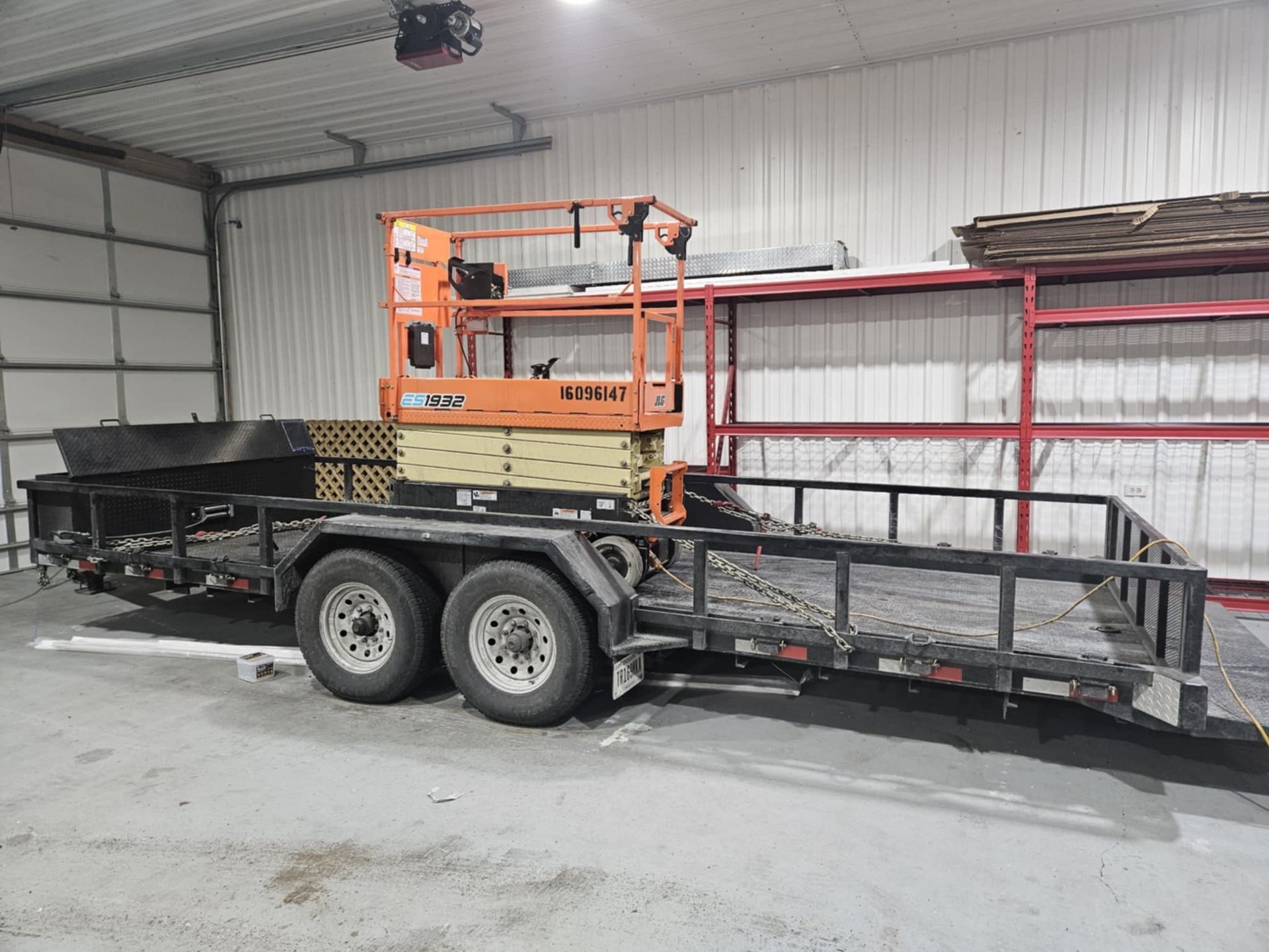 An orange and yellow scissor lift secured on a flatbed trailer parked inside a warehouse with tall storage shelving.