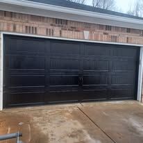A black, four-panel residential garage door on a brick home with a concrete driveway.