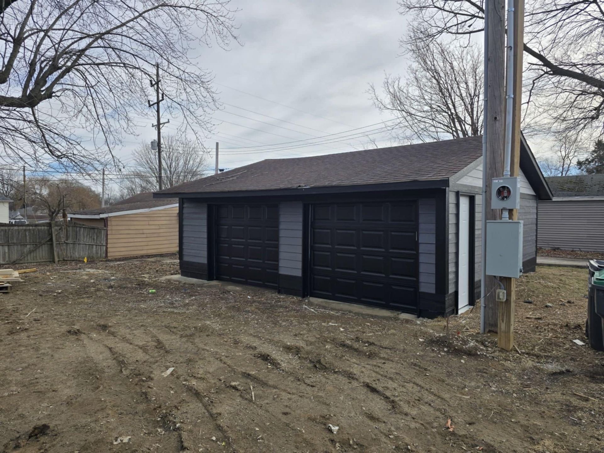 Newly built detached two-car garage with black doors and grey siding, set on an unfinished dirt lot.