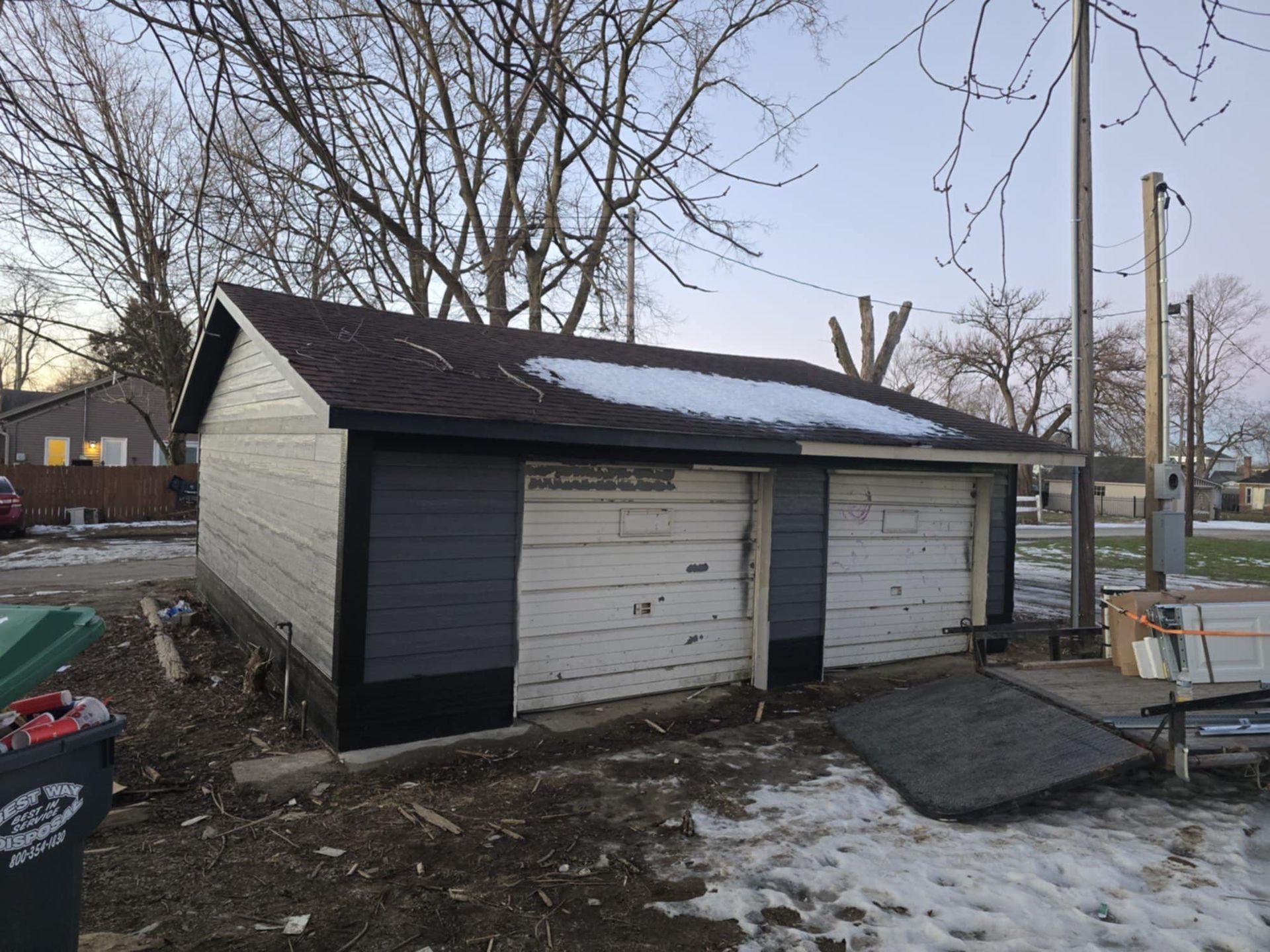 A detached garage with gray and white painted siding and two white garage doors under a roof with a patch of snow.
