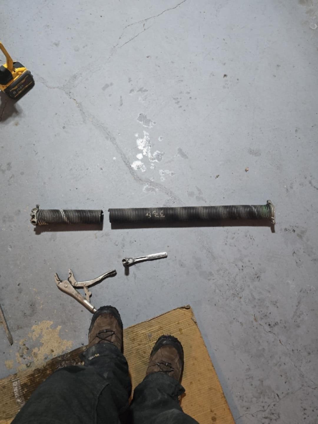 A broken metal garage door extension spring lies on a gray floor near tools and a person's boots.