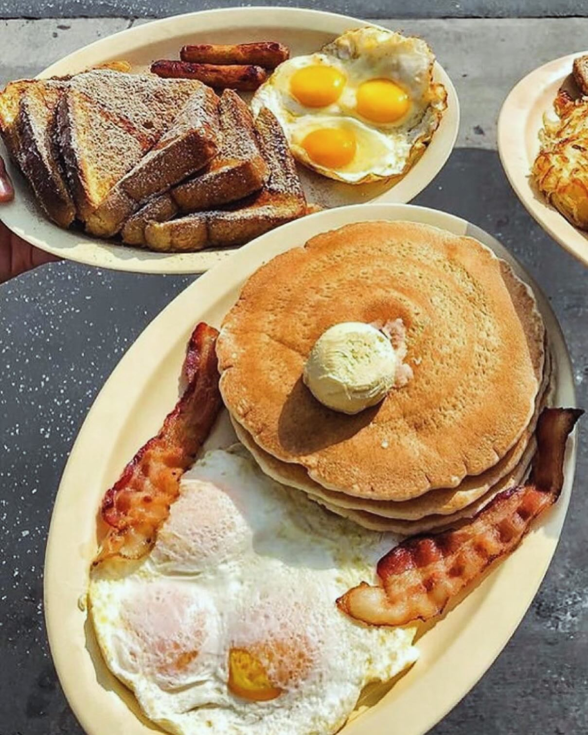 Plates of breakfast foods: pancakes with butter, fried eggs with bacon, french toast with sausage and eggs.