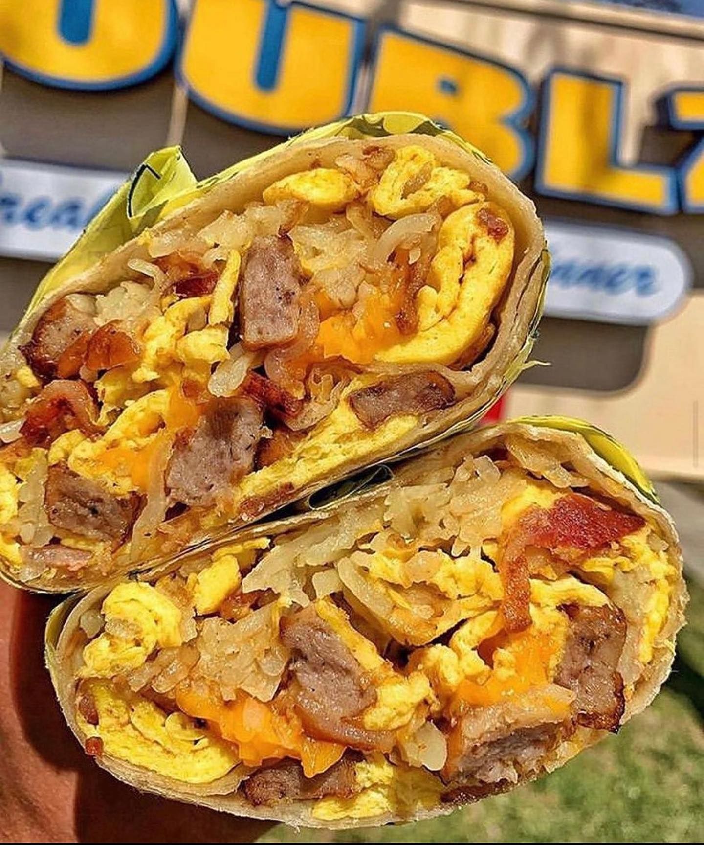Breakfast burrito cut in half, showcasing eggs, sausage, bacon, cheese, and hash browns. Yellow and orange tones with a yellow wrap.