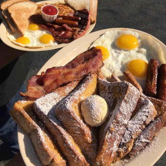Plates of breakfast food: fried eggs, bacon, sausage, toast, and french toast dusted with powdered sugar.