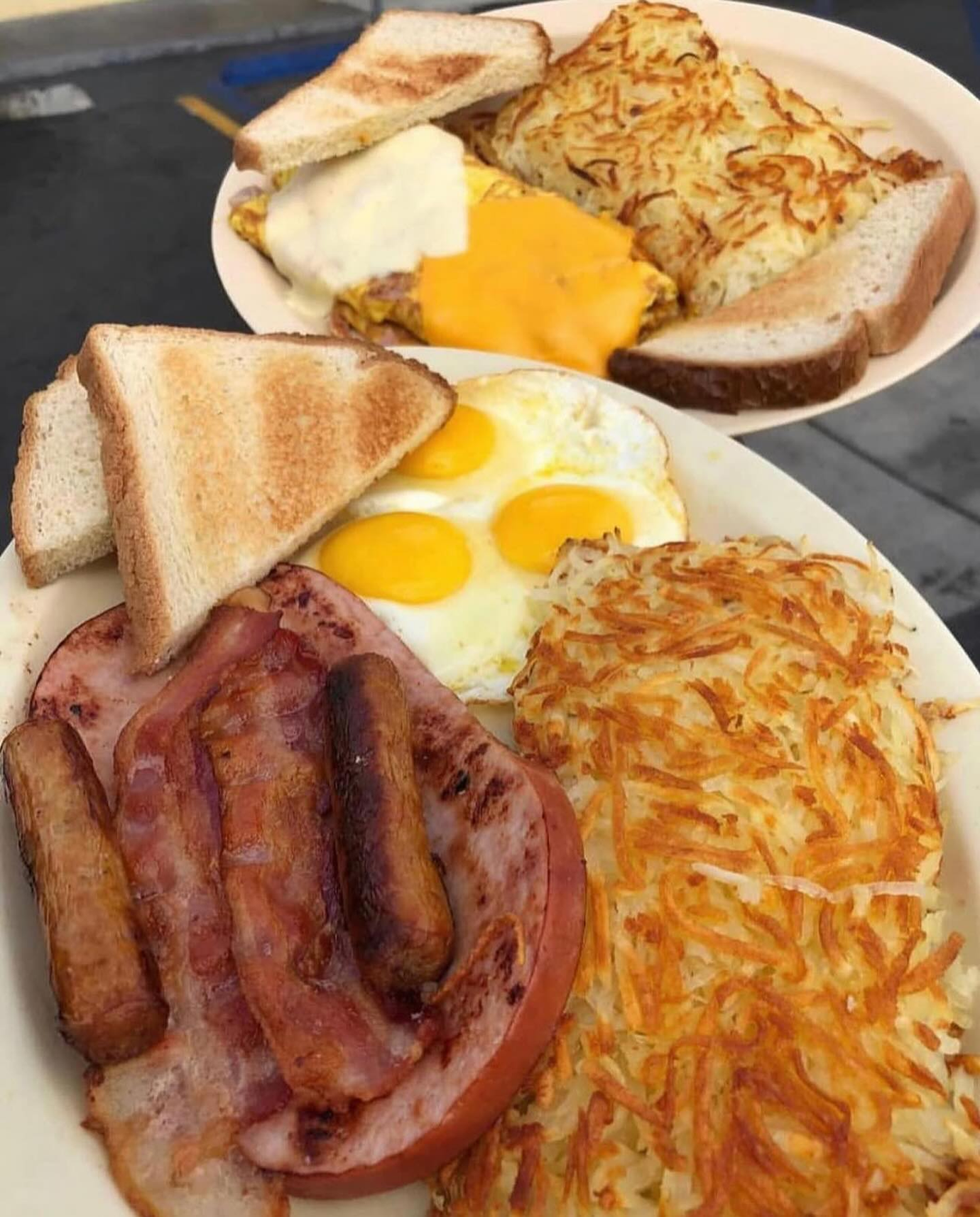 Two plates of breakfast food. One plate has eggs, hash browns, bacon, sausage and toast. The other has an omelet, hash browns, and toast.