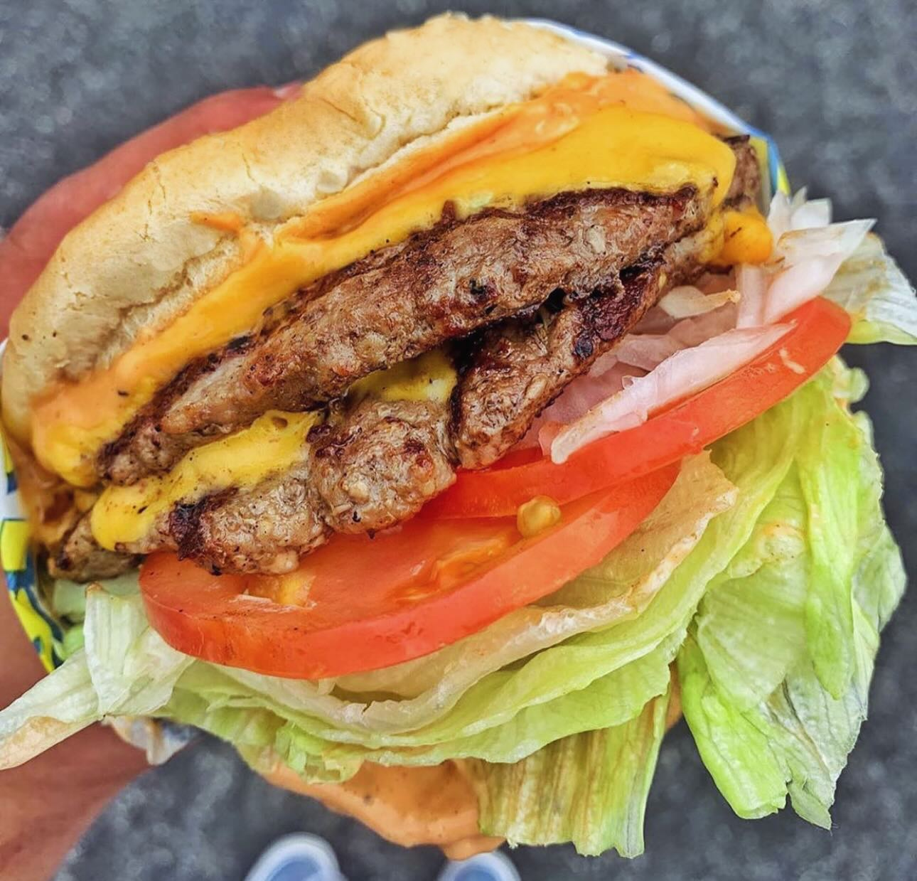 Double cheeseburger held in hand, showing beef patties, melted cheese, tomato, lettuce, and onion, on a sesame bun.