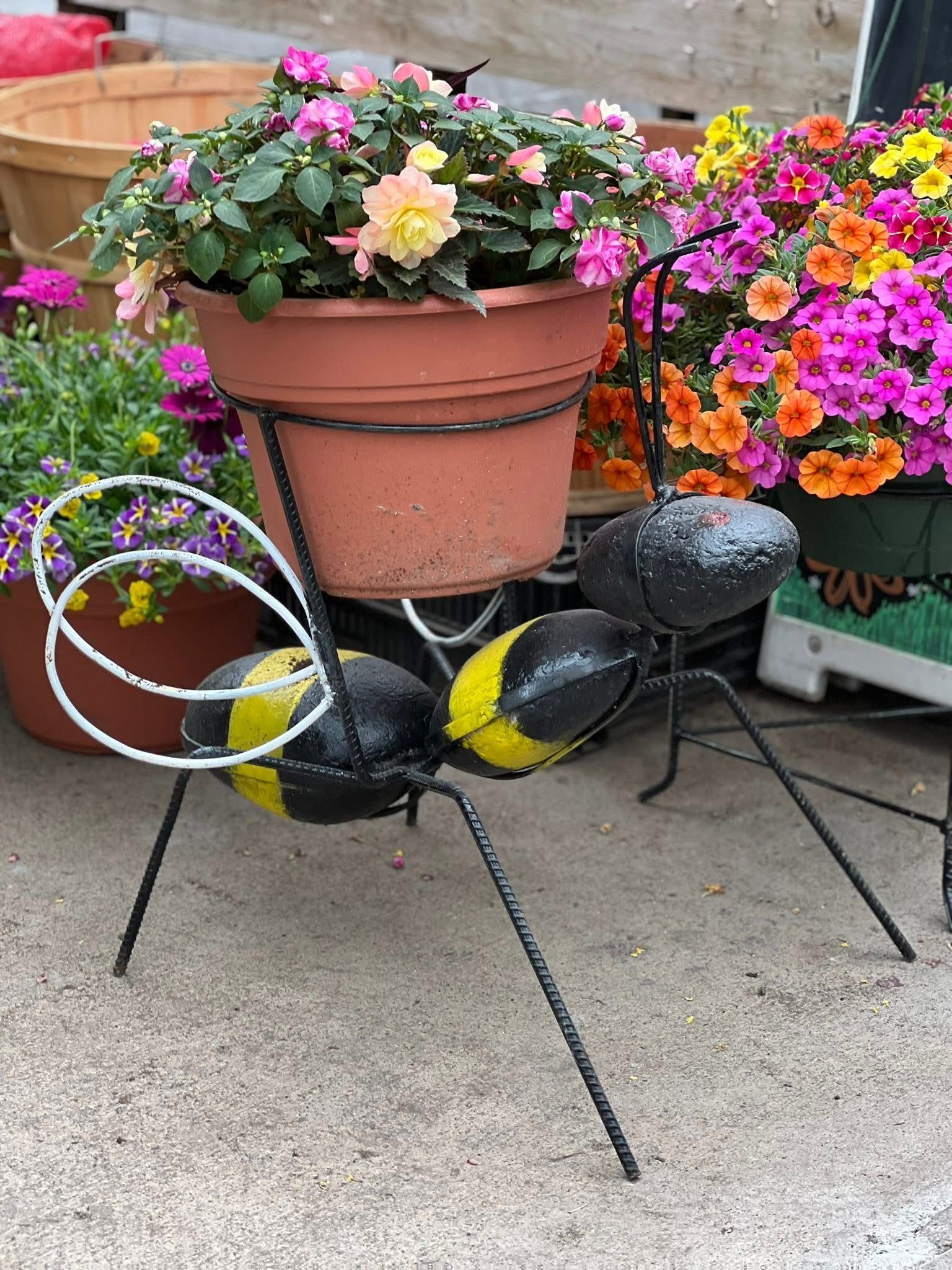 A novelty metal bee-shaped planter stand holding a terracotta pot filled with colorful pink and peach flowers.