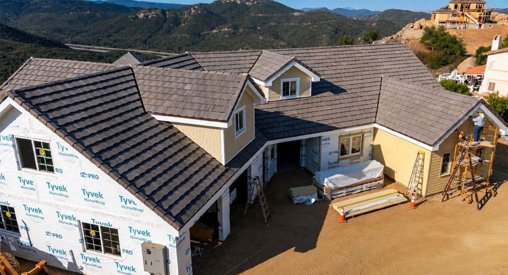 New house under construction with gray roof and white wrap, seen from above on a hillside lot