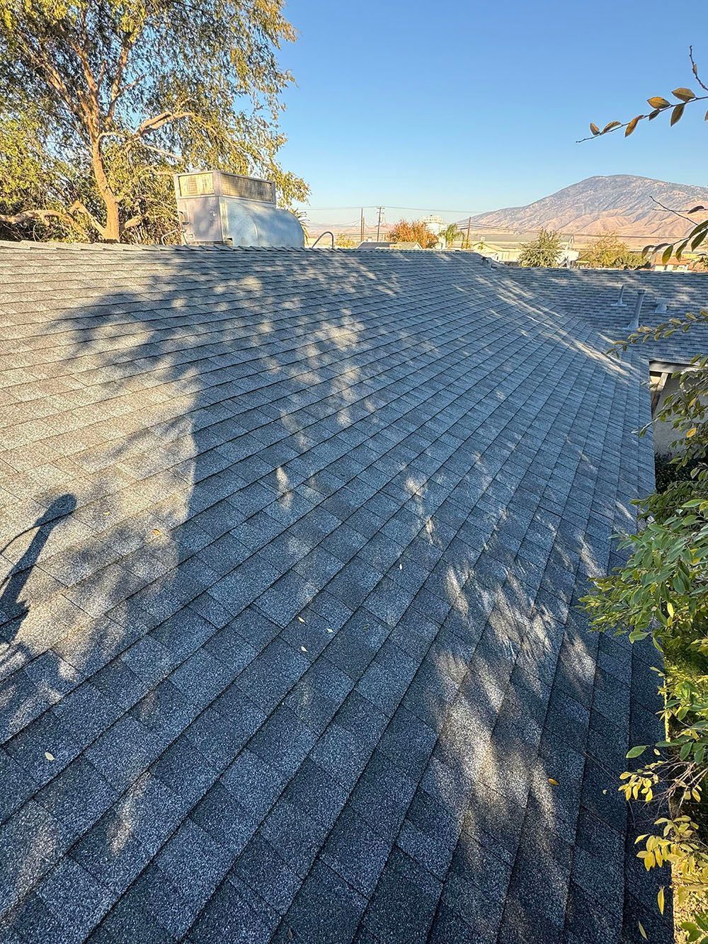 Steep gray shingle roof in sunlight with trees and mountains in the background