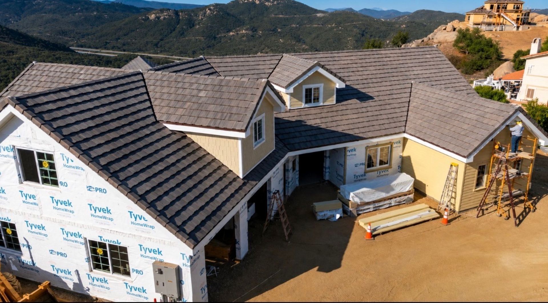 New home under construction with gray roof, wraparound porch, and wood framing on a hillside lot