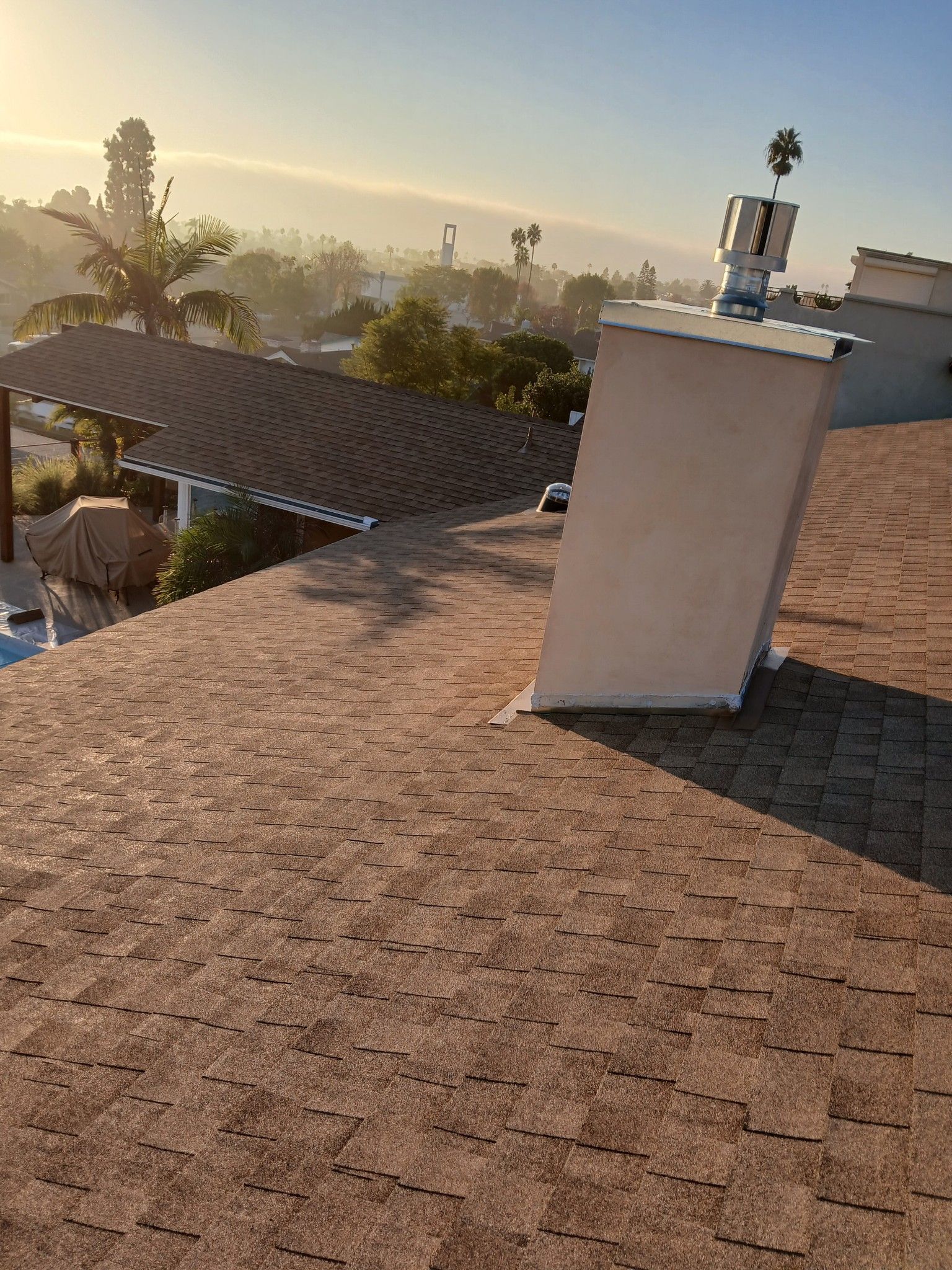 Rooftop at sunset with shingled roof, white vent, and palm trees in the distance.