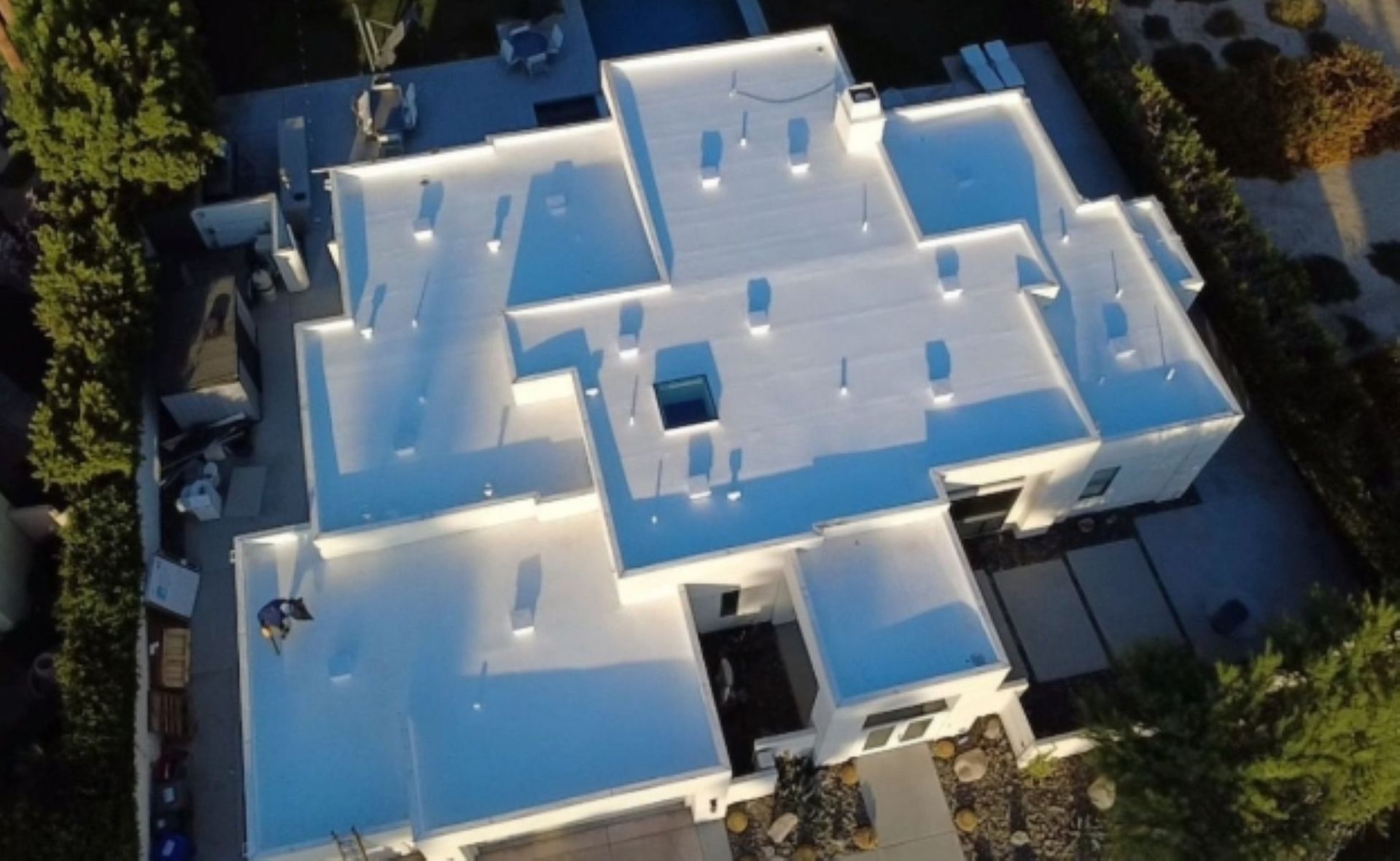 Aerial view of a white flat-roofed house with skylights, surrounded by trees and a driveway.