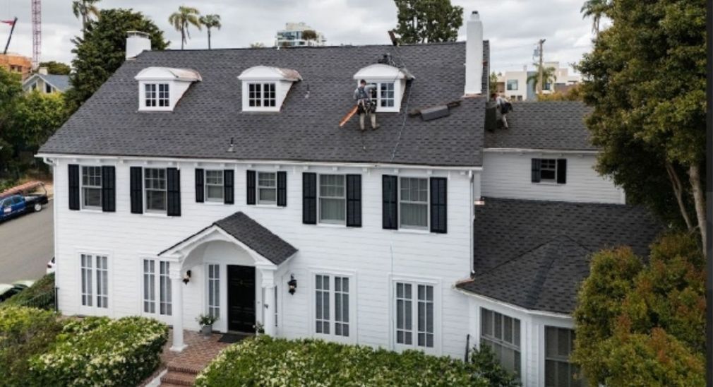 White two-story house with gray roof, dormer windows, black shutters, and a front entry surrounded by greenery
