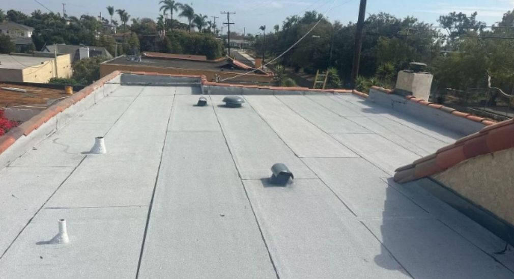 Flat rooftop with white waterproof coating and two vents, overlooking nearby trees and houses.