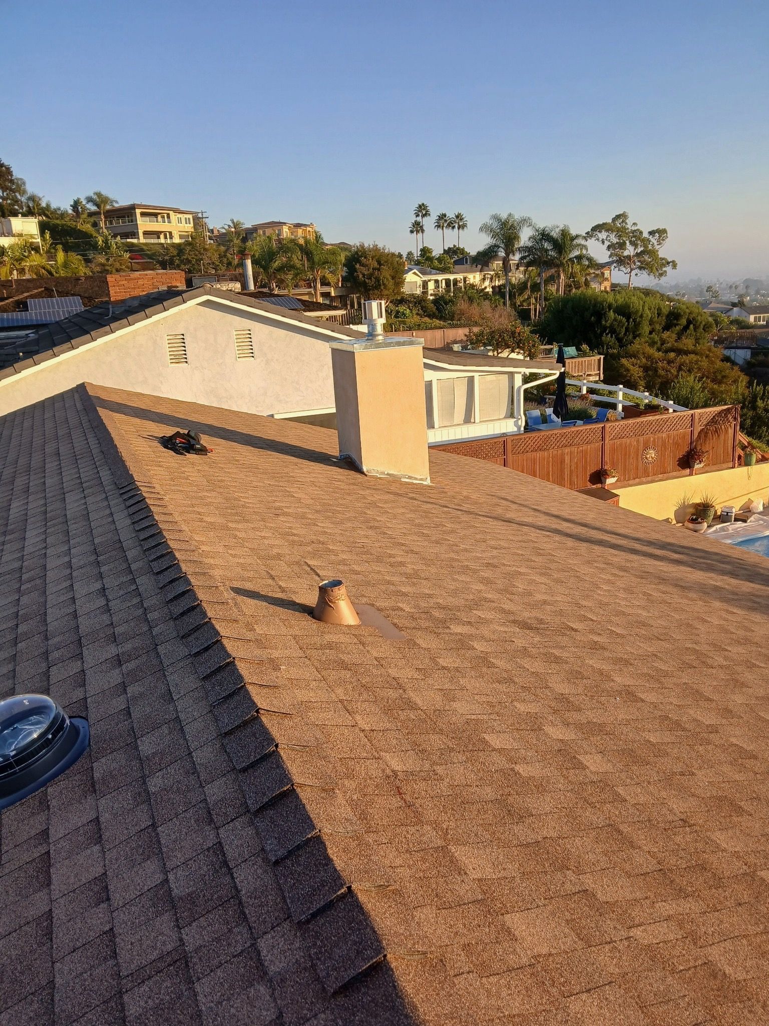 Rooftop view of a coastal neighborhood with tan shingles, palm trees, and hills in the distance