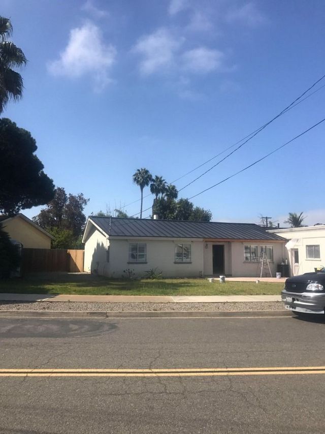 Single-story house with a tiled roof on a sunny street, with palm trees and a parked car nearby