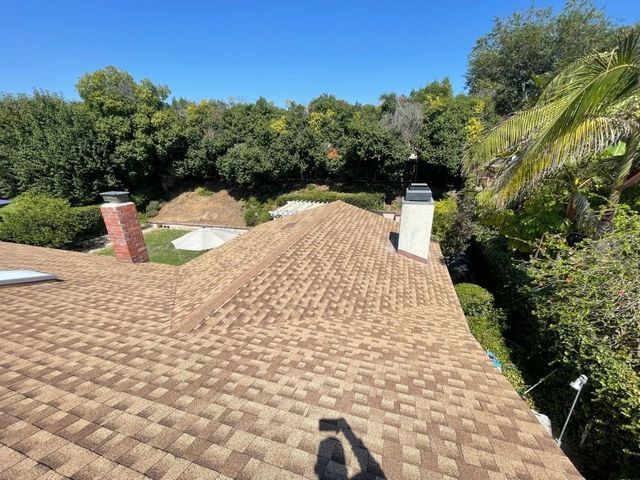 Rooftop covered in tan shingles with brick chimneys, surrounded by green trees under a clear blue sky