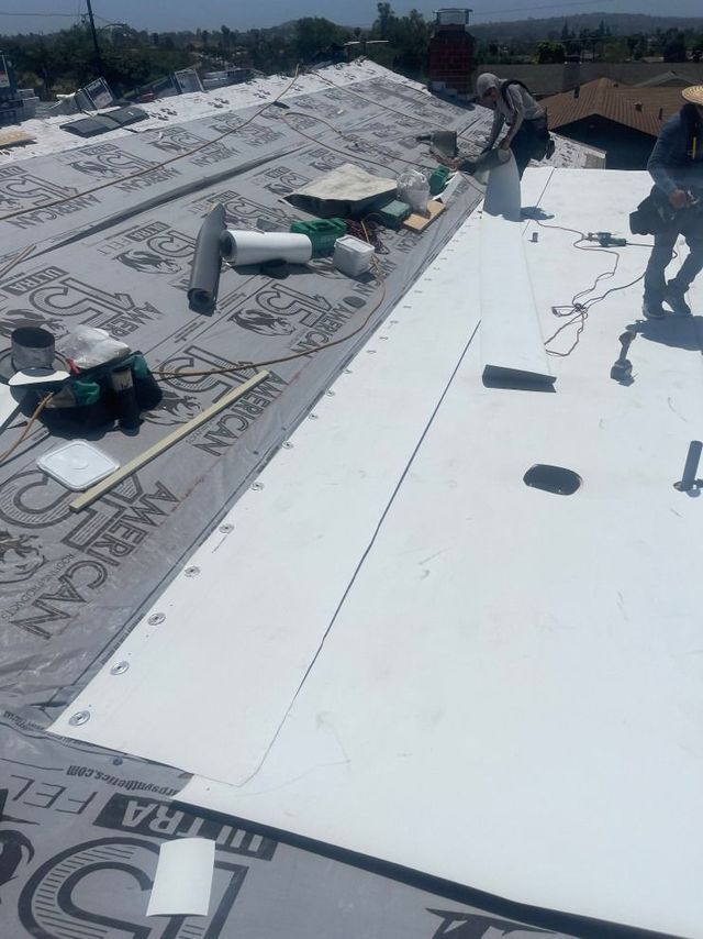 Workers installing a white roof membrane beside gray rooftop sections, with tools and materials spread out.