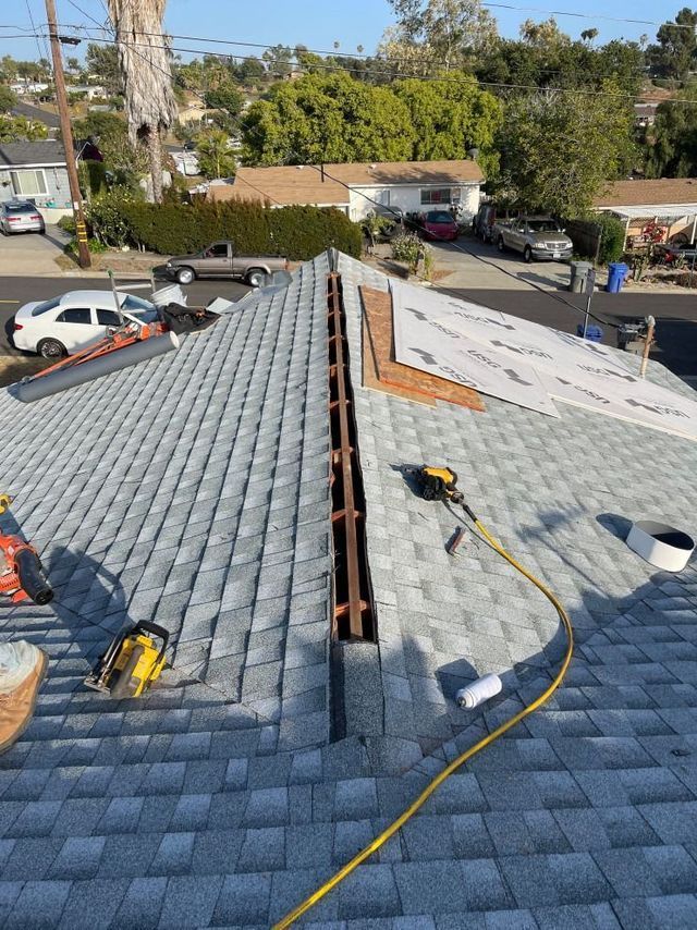 Roof repair with two workers on a gray shingle roof near a central ridge, tools and hoses visible