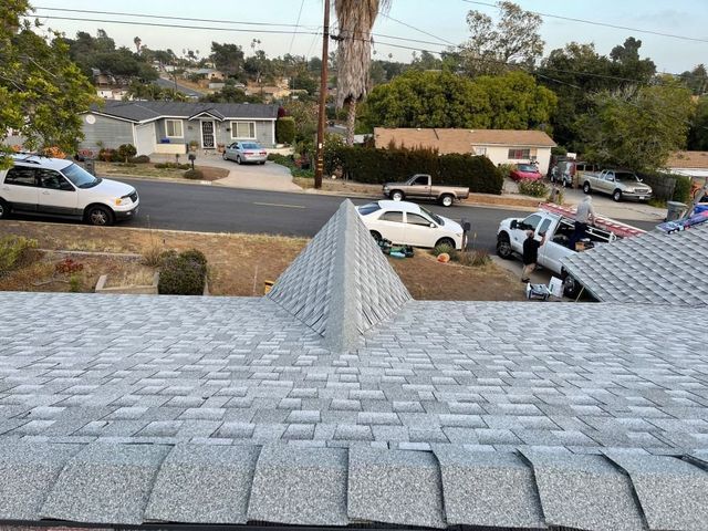 View from a shingled rooftop over a suburban street with parked cars and houses in the distance