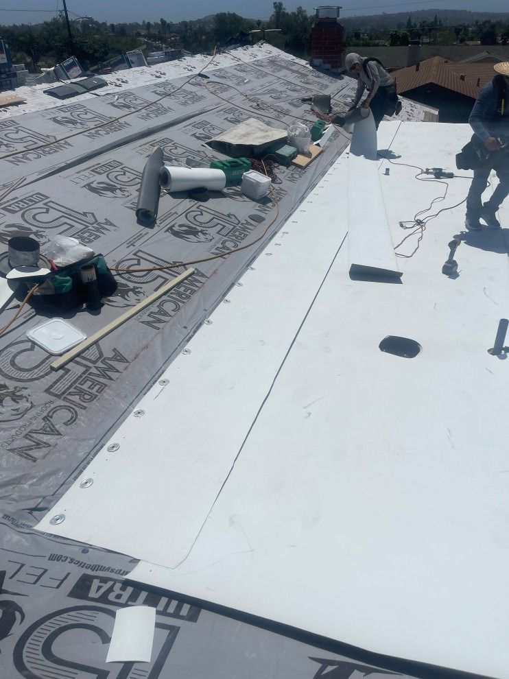 Workers installing white roofing material on a flat roof under a sunny sky