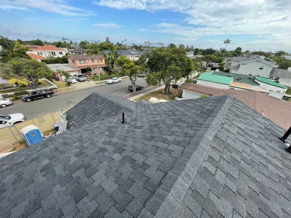 View from rooftop over suburban neighborhood with gray shingles, trees, houses, and a partly cloudy sky