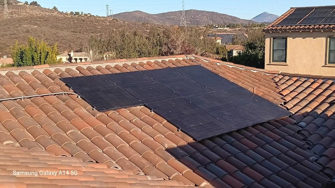 Sunlit terracotta rooftops with a large dark solar panel, overlooking dry hills and houses