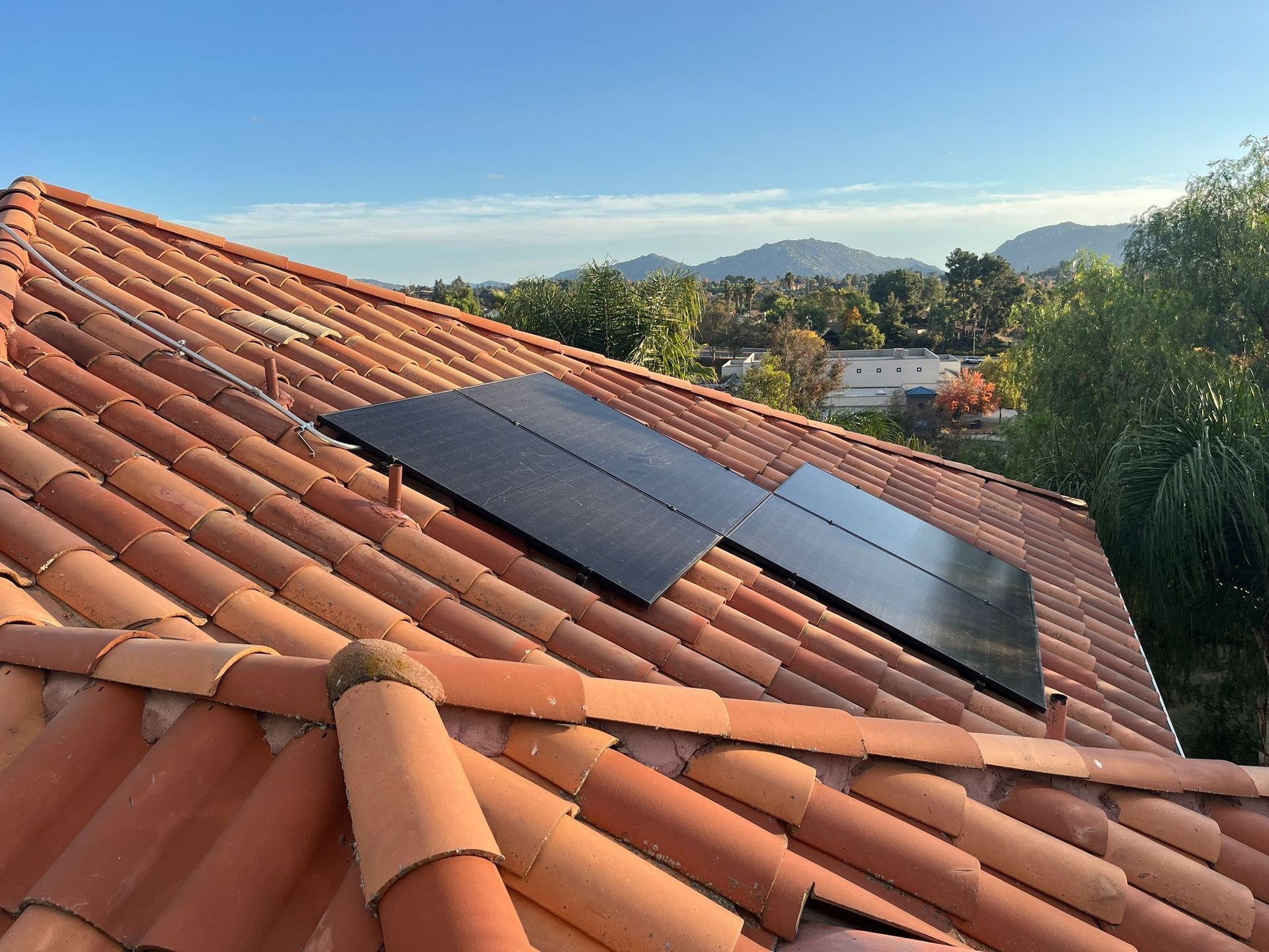 Black solar panels on a terracotta-tiled roof with trees and mountains in the background