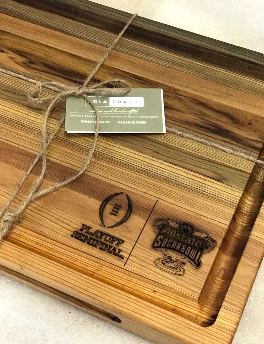 Wooden cutting board with engraved logos and a hanging tag on a striped wood surface