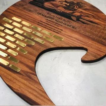 Wooden trophy plaque with gold nameplates and a large white cutout, engraved with a logo at the top.