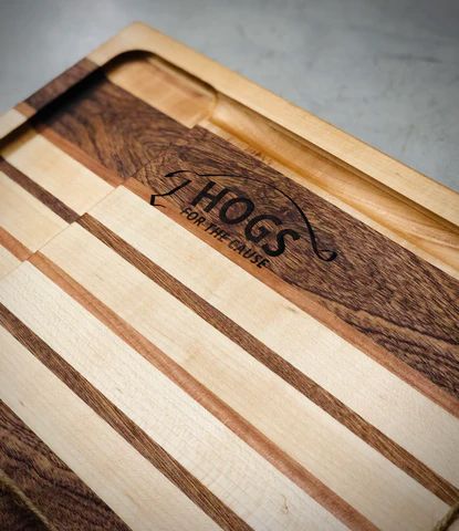Wooden cutting board with striped maple and dark walnut inlays and a carved logo near the top.