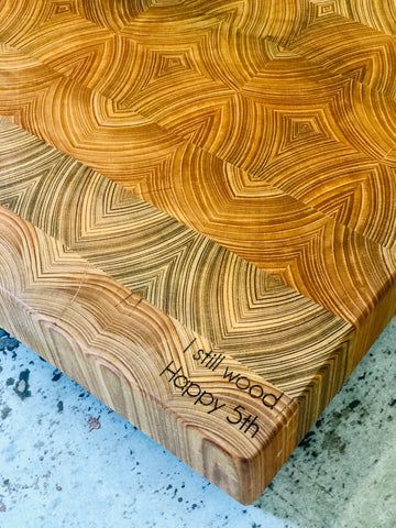 Wood end-grain cutting board with engraved text, in warm and light wood tones on a speckled countertop.