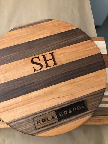 Round wooden board with dark stripes and initials 'SH' engraved, labeled 'NOLA BOARDS' at the bottom.