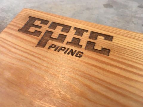 Wooden plaque with engraved black 'Epic Piping' logo on a light wood grain surface