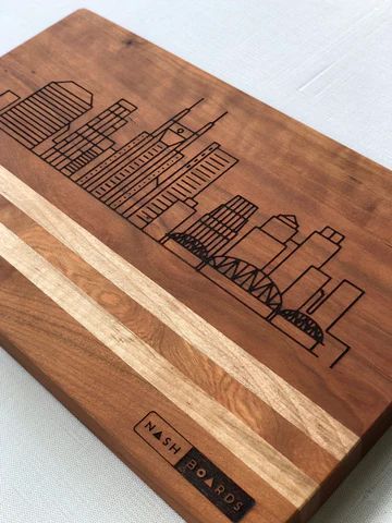 Laser-engraved city skyline on a wooden box lid with two light wood stripes and a small logo near the bottom