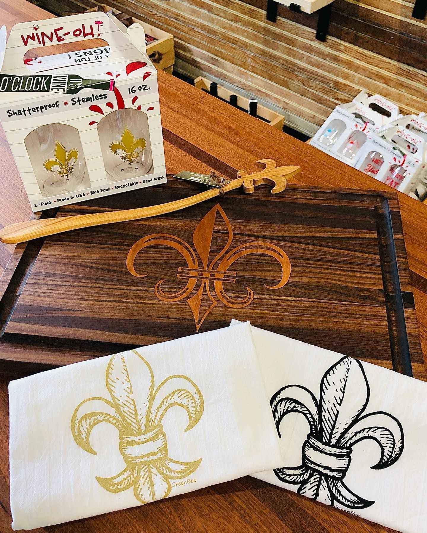 A wooden cutting board with a fleur-de-lis, matching towels, and a boxed set of wine glasses featuring the same emblem.