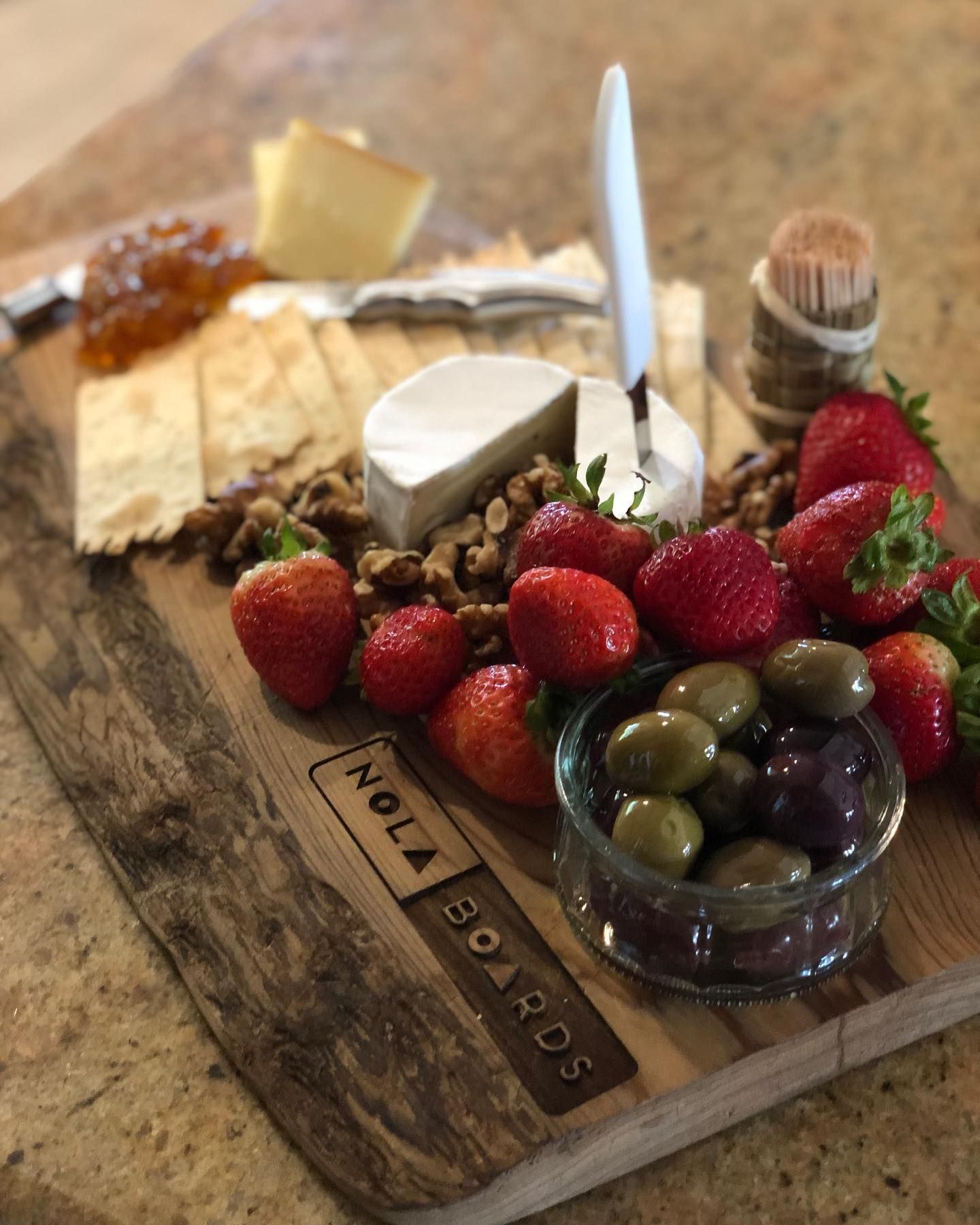 A wooden Nola Boards charcuterie board with cheese, crackers, strawberries, walnuts, olives, and a serving knife.