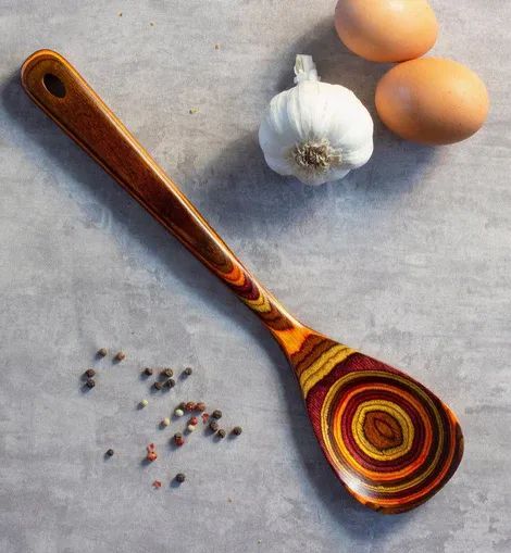 A multicolored wooden cooking spoon rests on a gray surface next to two eggs, a head of garlic, and scattered peppercorns.