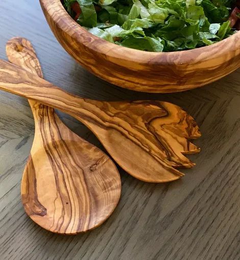 A wooden salad bowl filled with lettuce, next to two decorative olive wood salad serving utensils on a gray surface.
