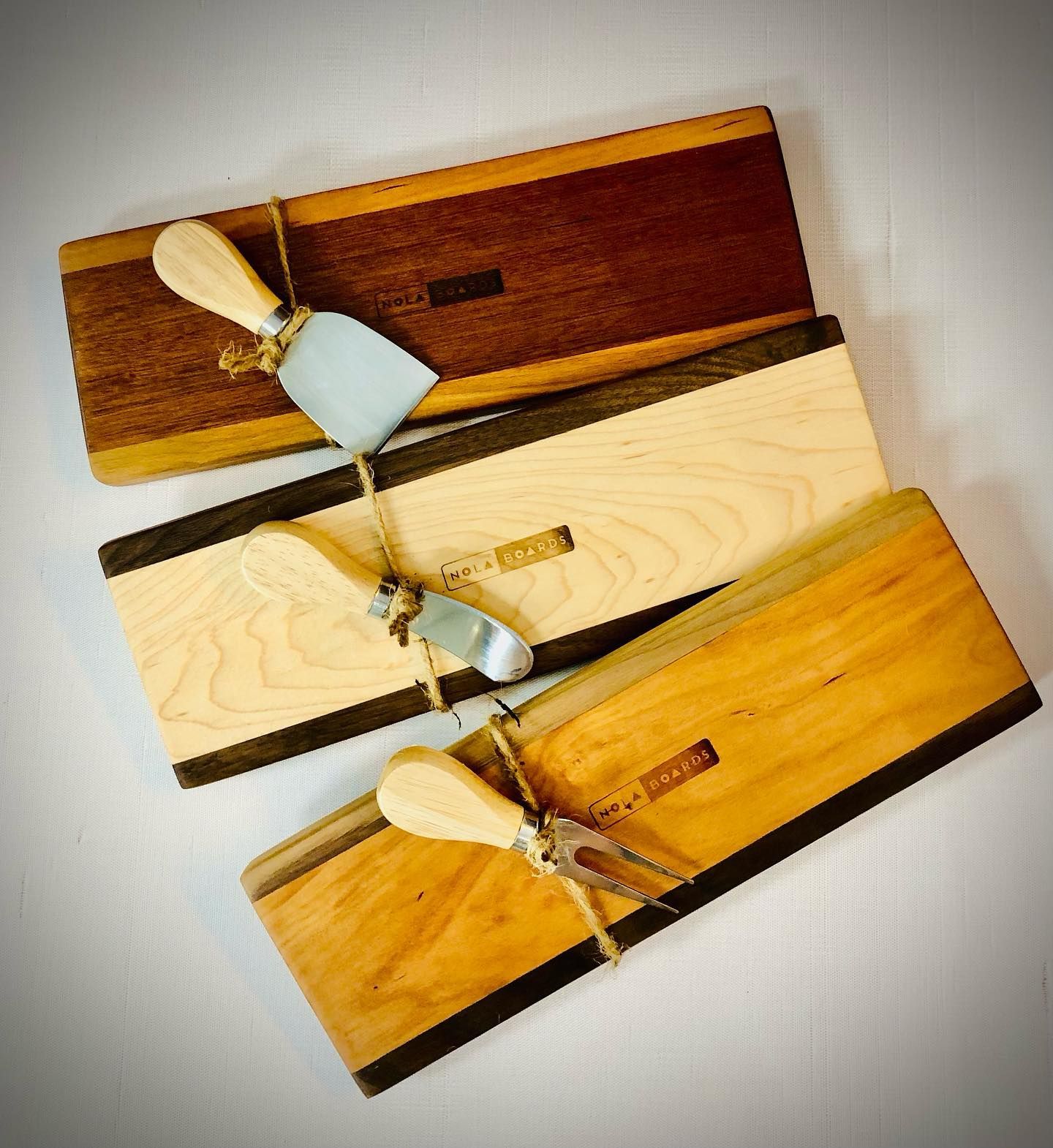 Three rectangular wooden cheese boards with matching serving utensils tied by twine on a white background.