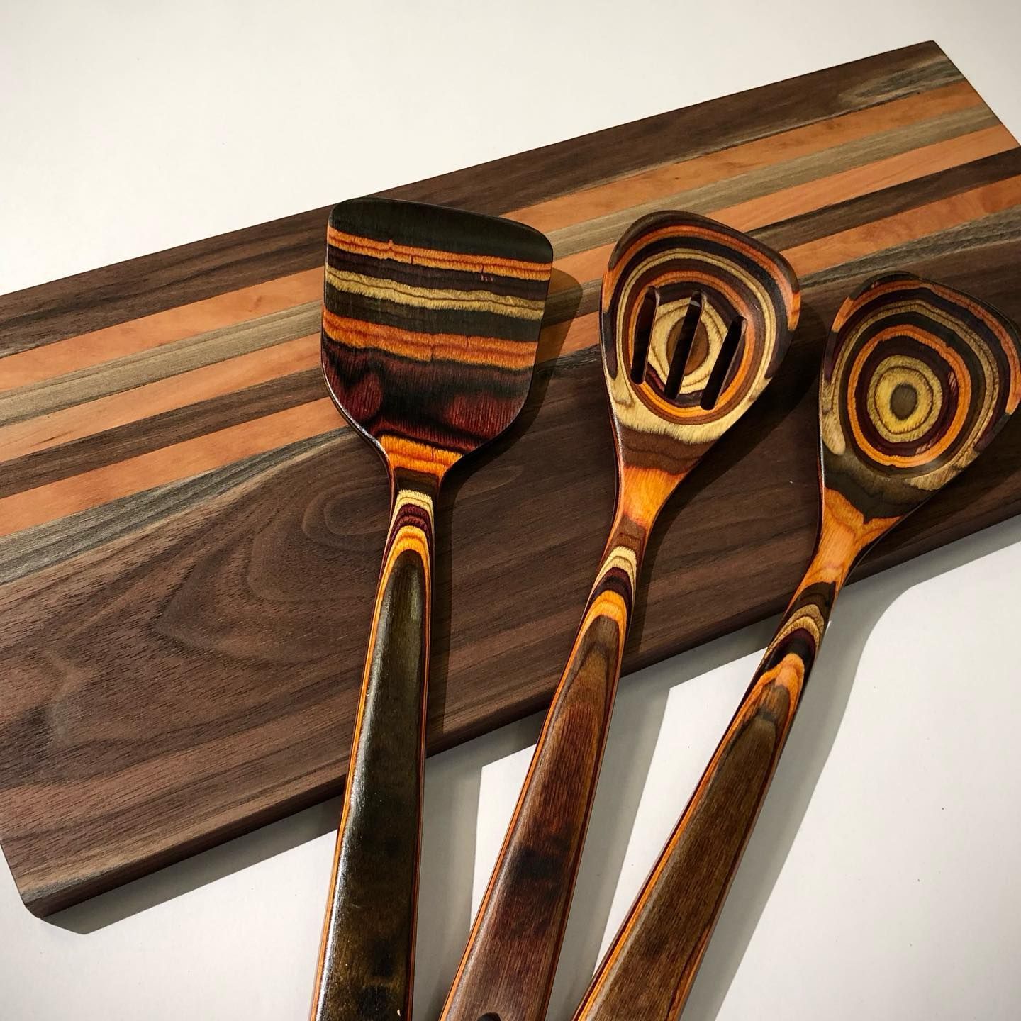 A wooden cutting board with distinct stripes sits behind a set of three matching, multi-colored, laminated wooden utensils.