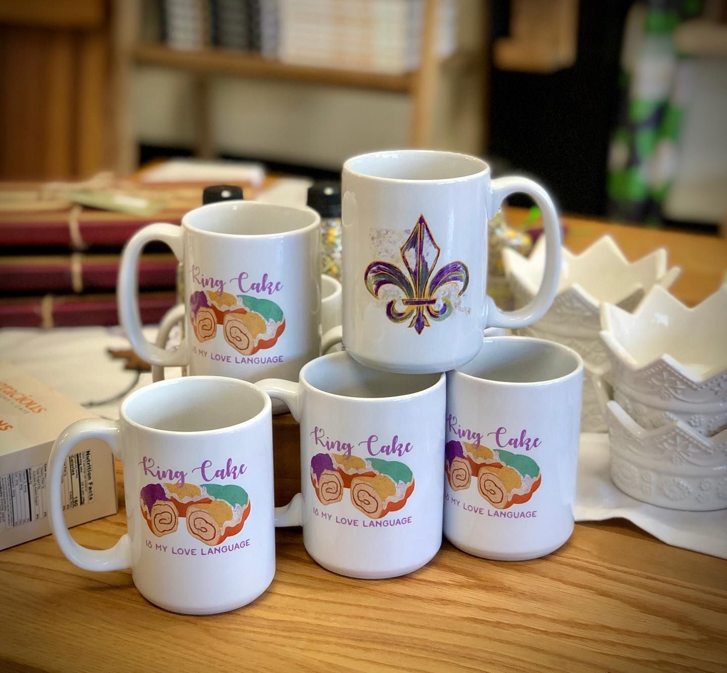 Five white mugs displayed on a wooden surface, four featuring a King Cake design and one showing a purple and gold fleur-de-lis.
