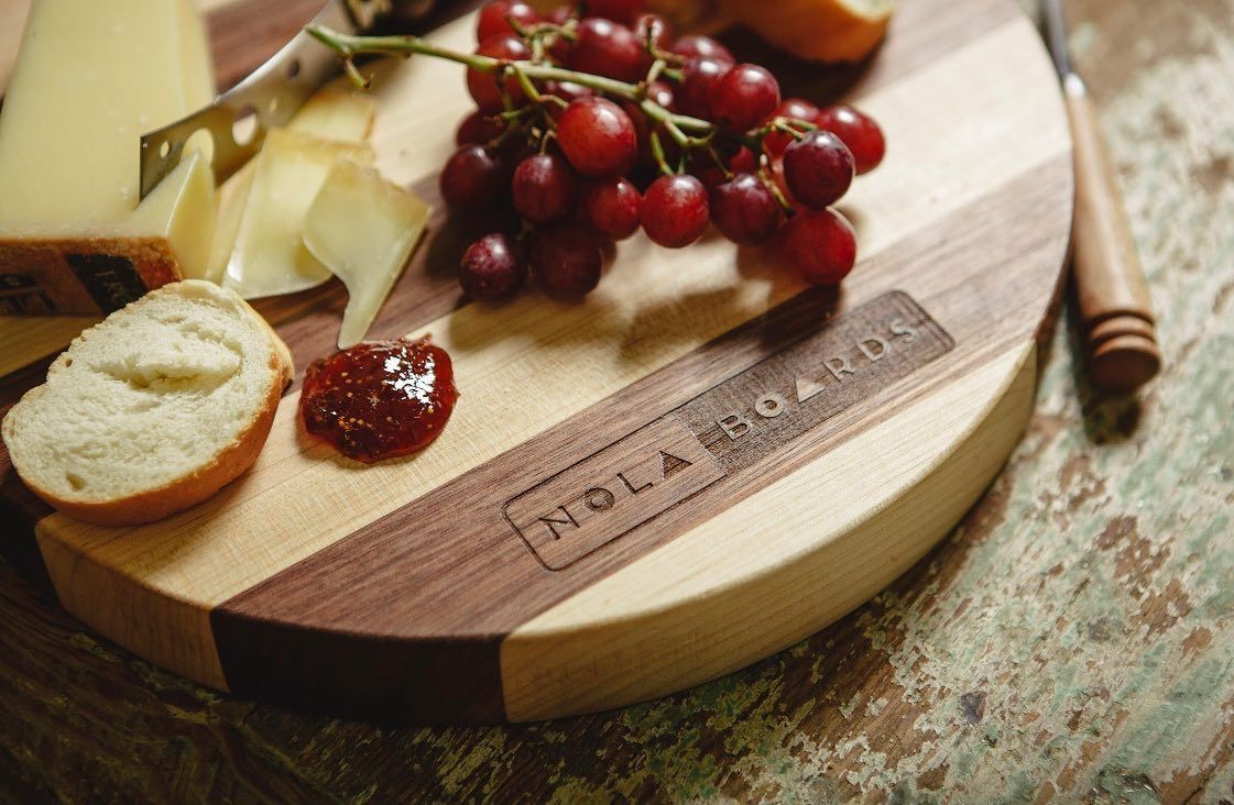 A charcuterie board featuring grapes, cheese, a piece of bread, and jam, with a NOLA Boards logo on the wood surface.