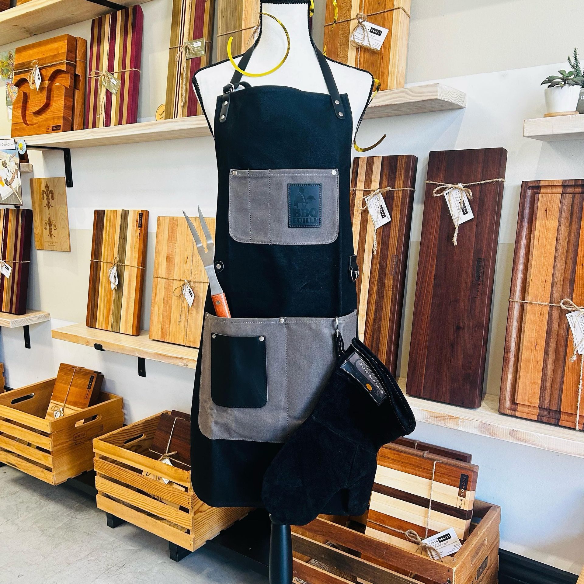 A black and gray apron with pockets and a grill fork displayed on a mannequin in a store stocked with wooden boards.